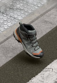 Szare buty trekkingowe z syntetyczną cholewką, gumową podeszwą, pomarańczowymi akcentami i zintegrowanymi sznurowadłami. Posiadają wyściełany kołnierz dla wsparcia.