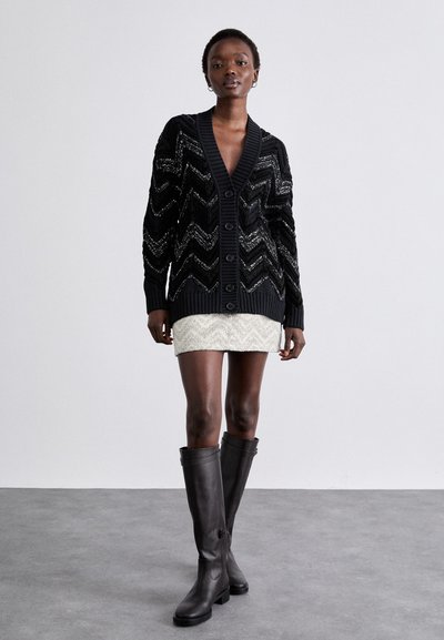 Μαύρο κασκόλ με σχέδιο ζιγκ-ζαγκ και ασημένιες λεπτομέρειες, με κουμπιά, ριμπ άκρη και μανίκια, φορεμένο πάνω από μια καρό κρέμα φούστα και ψηλές καφέ μπότες.