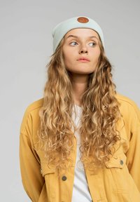 Jasnoniebieska, prążkowana czapka z podwijanym brązowym skórzanym naszywkiem. Noszona z żółtą kurtką i białą koszulą, z długimi, kręconymi włosami.