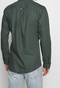 Camisa de cuadros verde y azul marino con mangas largas, puños abotonados, dobladillo curvado y etiqueta en la parte trasera. Combinada con vaqueros azul claro.