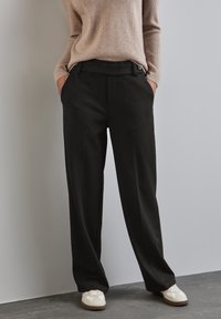 Pantaloni neri su misura con una vestibilità rilassata, dotati di tasche laterali e una superficie liscia. Abbinati a un maglione beige e sneakers bianche.