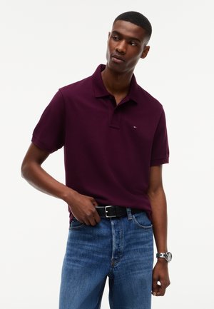 Joven que lleva una camiseta tipo polo de color púrpura oscuro y vaqueros azules, ajustando un cinturón negro, con un reloj de pulsera plateado en su muñeca izquierda.