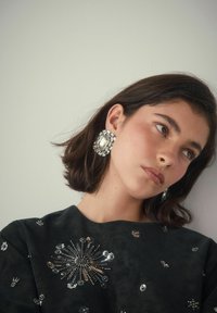 Junge Frau mit kurzen dunklen Haaren, die große, klare Edelstein-Ohrringe und ein schwarzes Oberteil mit silbernen Paillettenmustern trägt, blickt seitlich.