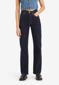 Femme portant un jean droit taille haute bleu foncé, des chaussures noires sans lacets et une veste en jean courte sans manches, debout avec une main dans la poche.