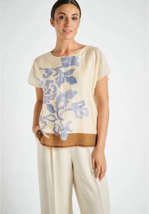 Vrouw draagt een beige blouse met korte mouwen, blauw bloemmotief en een bruine zoom, gecombineerd met losse crèmekleurige broek, staande tegen een witte achtergrond.