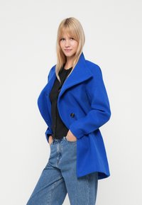Cappotto di lana blu con ampio colletto, chiusura doppio petto e grandi bottoni neri. Abbinato a un top nero e jeans azzurri chiari.
