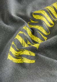 Szürke szövet puha textúrával és kopott sárga grafikai mintával, hullámos vonalakkal különböző vastagságban.