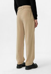 Beige broeken met een rechte snit, voorzien van een tailleband, twee achterzakken, en gedragen met zwarte schoenen, die een gladde textuur tonen.