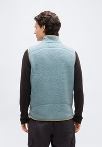 Άνδρας που φοράει ανοιχτό μπλε γιλέκο fleece με κίτρινη επένδυση πάνω από μαύρο μακρυμάνικο μπλουζάκι, εμφανιζόμενος από πίσω, σε απλό φόντο.