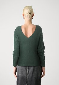 Zelený pletený svetr s hloubokým výstřihem na zádech do V, dlouhými rukávy a žebrovanými manžetami. Nošený přes šedou džínovou sukni. Textura působí robustně a útulně.