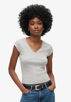 Top grigio chiaro a maniche corte con scollatura a V, texture a costine e orlo in pizzo ondulato lungo il collo, abbinato a jeans in denim scuro e una cintura nera.