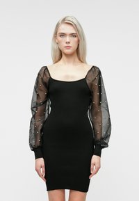 Mulher loira vestindo um vestido preto justo com mangas bufantes transparentes adornadas com pequenas pérolas, de pé contra um fundo branco simples.