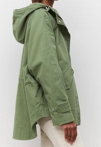 Zielona, lekka kurtka z kapturem, z bocznymi kieszeniami i guzikiem zatrzaskowym. Materiał jest gładki, o luźnym kroju i dłuższym tylnym hemie.
