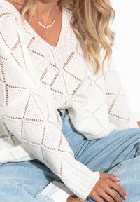 Weißer Strickpullover mit einem rautenförmigen Muster, tiefem V-Ausschnitt und langen Ärmeln, kombiniert mit hellblauen Jeans und weißen Schuhen.