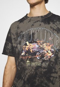 Tmavě šedé tričko s tie-dye vzorem, které má grafiku auta se plameny, text "COUTURE" nahoře a "THE COUTURE CLUB" dole ve zlaté barvě.