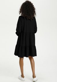Schwarzes, mehrstufiges Kleid aus weichem Stoff mit langen Ärmeln und ausgestellter Silhouette. Kombiniert mit weißen Turnschuhen für einen lässigen Look.