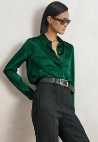 Frau trägt Schildpatt-Sonnenbrille, smaragdgrüne Satinbluse, schwarze Hose und schwarzen Gürtel mit goldener Schnalle und posiert vor einer einfarbigen Wand.