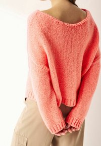 Różowy sweter robiony na drutach o luźnym kroju, szerokim dekolcie i ściągaczach. Miękka tekstura i swobodna sylwetka, połączone z beżowymi spodniami.