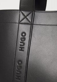 Sac fourre-tout en cuir noir avec logo "HUGO" en relief. Texture lisse avec couture visible et détails de surpiqûre sur la poignée.