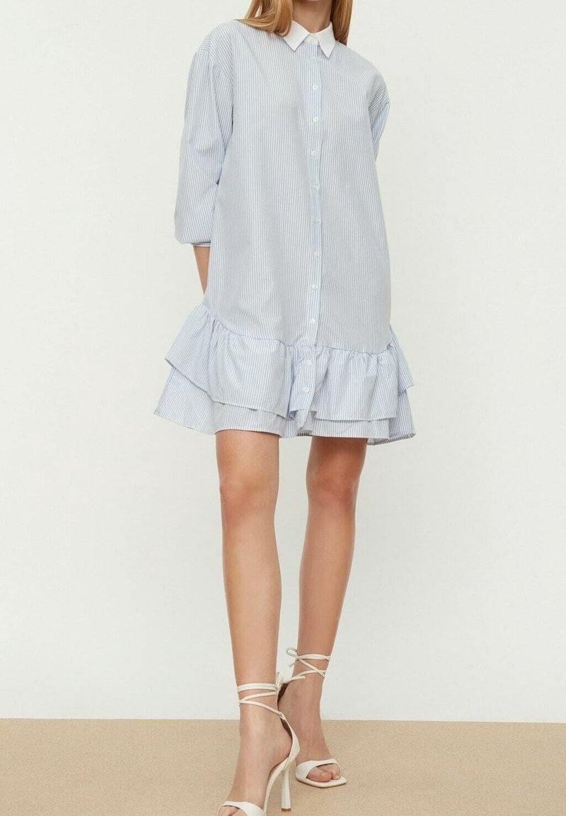 Femme portant une robe chemise rayée bleu clair avec un ourlet volanté et des sandales blanches à talons hauts nouées à la cheville, debout devant un fond uni.