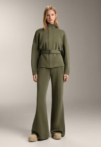 Olive zelená súprava na voľný čas s opaskom a širokými nohavicami, vyrobená z mäkkej tkaniny; má vysoký golier a nadrozmerný strih.