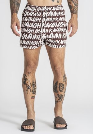 Homme portant un short marron et blanc à motifs avec le texte répété « GIANNI KAVANAGH », debout avec des tatouages visibles sur les jambes et les bras, et des sandales marron à enfiler.