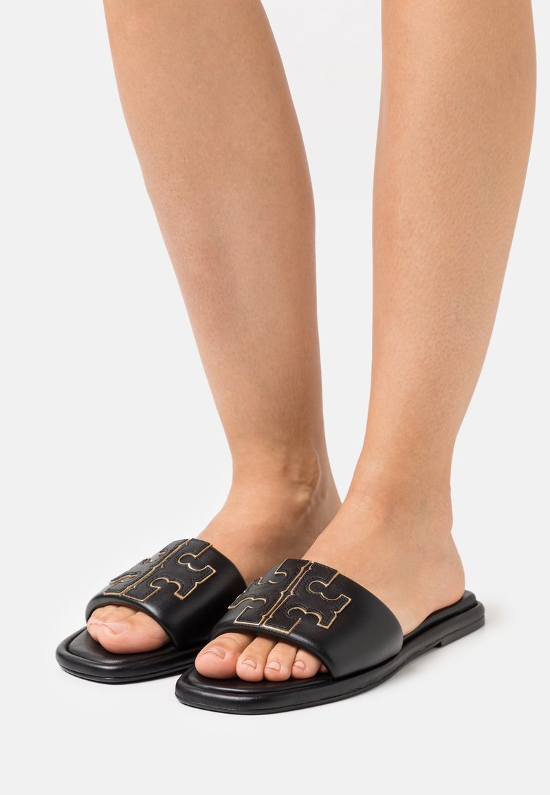 Pieds portant des sandales noires à brides larges avec un logo découpé symétrique cousu, sur un fond blanc.