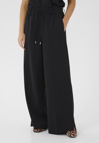 Čierne nohavice s širokými nohavicami, elastickým pásom, so šnúrkou, bočnými vreckami a hladkou textúrou. Viditeľné sú topánky s leopardím vzorom.