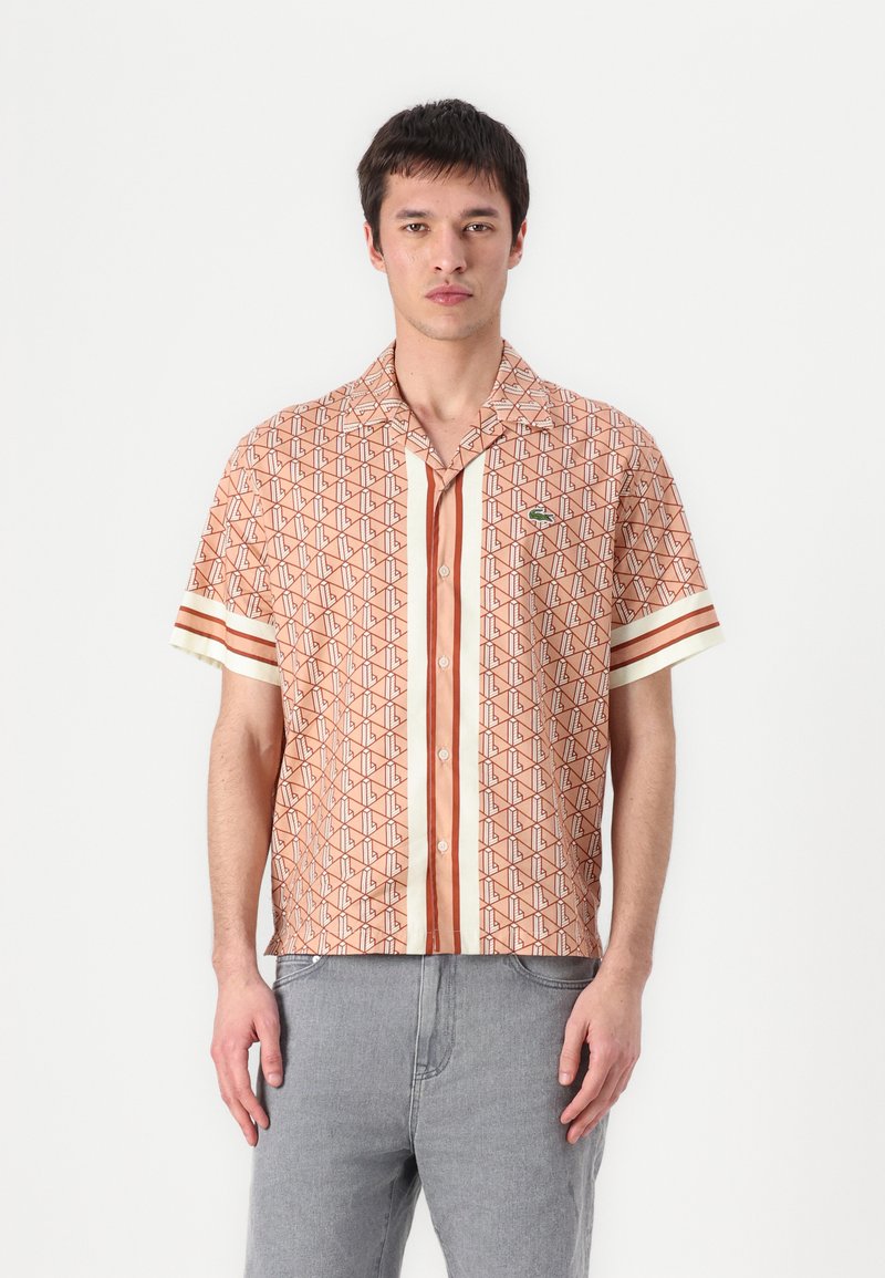 Kortermet skjorte i oransje mønstret stoff med geometriske design, horisontale striper og en liten grønn logo på venstre bryst.