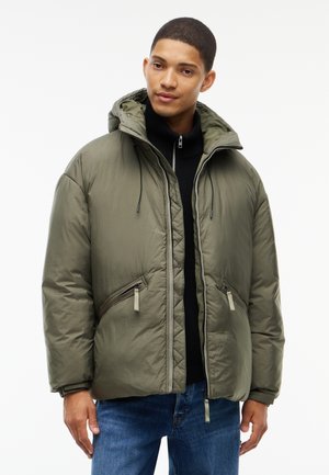 Jeune homme portant une veste matelassée à capuche vert olive par-dessus un pull noir zippé et un jean bleu, debout devant un fond blanc.