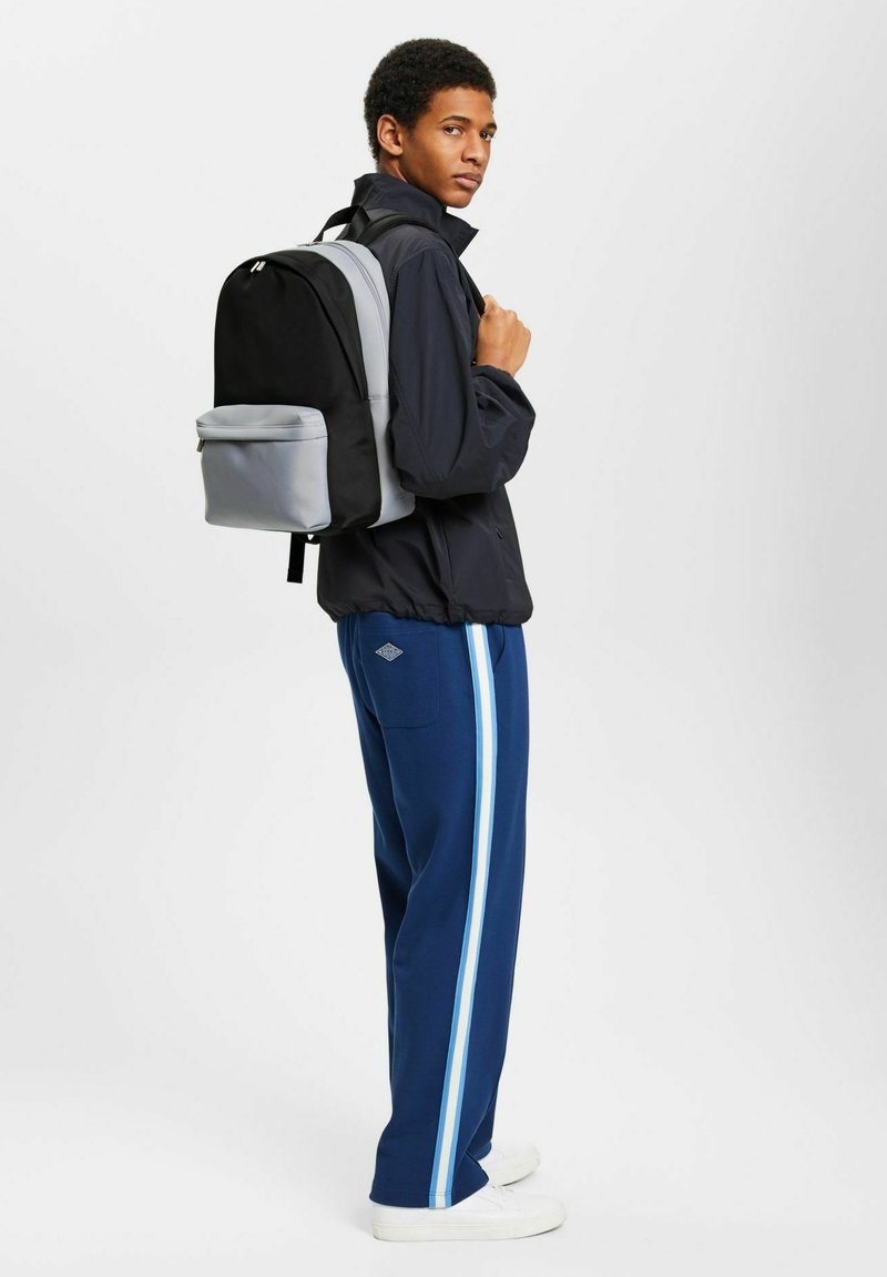 Mochila negra y gris con un diseño estructurado, que cuenta con correas ajustables y un bolsillo frontal con cremallera. La lleva una persona que viste pantalones deportivos.