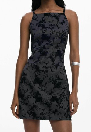 Femme portant une robe sans manches noire et grise à motif abstrait avec des bretelles fines et un bracelet manchette en argent sur l'avant-bras gauche.