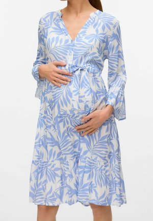 Kobieta w ciąży w niebiesko-białej sukience w liście, delikatnie obejmująca brzuch obiema rękami na jednolitym tle.