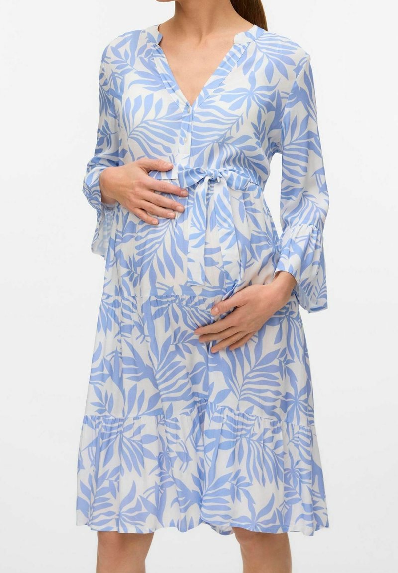 Femme enceinte vêtue d'une robe bleue et blanche à motif de feuilles, tenant délicatement son ventre avec les deux mains sur un fond uni.