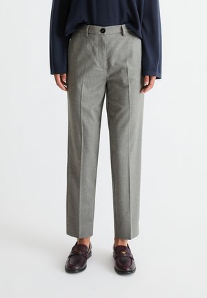 Personne portant un pantalon gris ajusté, des mocassins foncés et un haut à manches longues bleu marine, debout devant un fond blanc uni.