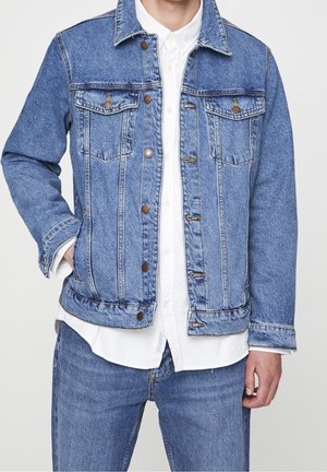 Mann trägt blaue Jeansjacke über weißem Hemd mit Knöpfen und passende blaue Jeans, steht mit einer Hand in der Tasche.