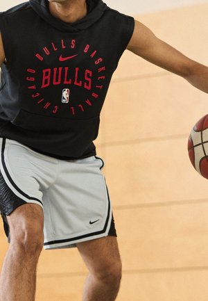 Un joueur de basketball masculin porte un hoodie sans manches des Chicago Bulls et un short blanc de Nike, dribbler un ballon de basketball à l'intérieur.