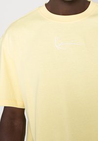 Gul bomullströja med rund halsringning, prydd med en vit broderad signatur på bröstet. Slät textur och avslappnad passform.