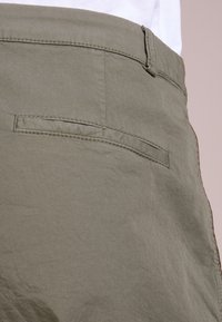 Pantaloni corti in tessuto verde oliva con una texture liscia, dotati di una tasca posteriore e passanti per cintura. Il capo ha una leggera lucentezza e cuciture nette.