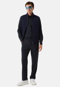 Granatowa pikowana kamizelka na ciemnej koszuli z guzikami, w połączeniu z dopasowanymi spodniami i białymi sneakersami. Duże niebieskie okulary przeciwsłoneczne uzupełniają stylizację.