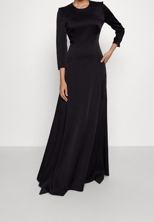 Woman wearing a long black satin dress with three-quarter sleeves, fitted bodice, and a flowing skirt standing against a plain background.