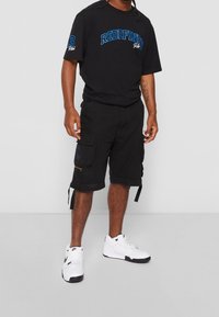 Mężczyzna ubrany w czarną koszulkę z napisem "Redefined", czarne szorty cargo z kieszeniami i białe trampki Nike, stoi na jednolitym tle.