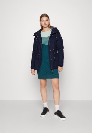 Giacca parka blu navy con cappuccio, dotata di zip e chiusura con coulisse, indossata sopra un vestito turchese, abbinata a sneakers nere.