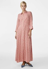 Femme portant une longue robe fleurie rose boutonnée à manches trois-quarts et des escarpins bleus à bout pointu, se tenant devant un fond uni.