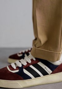 Baskets en suède rouge et marine avec des rayures blanches, dotées d'un dessus texturé et d'une semelle en caoutchouc. Pantalon beige avec un revers visible au-dessus des chaussures.