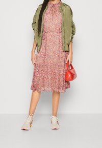 Veste bomber en satin vert, robe midi florale aux tons roses et violets, sac à main rouge et chaussures de sport beiges avec des accents roses.