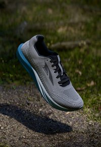 Chaussure de sport grise avec une tige en mesh, lacets noirs et une semelle bleu clair. Présente un logo sur le côté et un col de cheville rembourré.