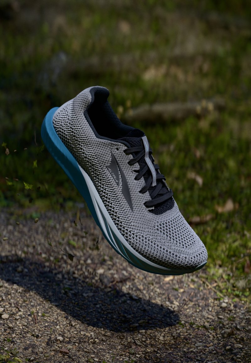 Chaussure de sport grise avec une tige en mesh, lacets noirs et une semelle bleu clair. Présente un logo sur le côté et un col de cheville rembourré.