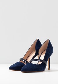 Un paio di décolleté in suede blu navy con punta affusolata e una brillante fascia nera sulla parte superiore, disposte su uno sfondo bianco semplice.