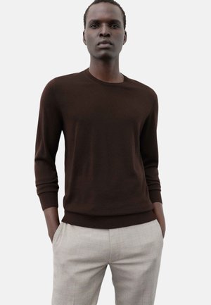 Man wearing a dark brown long-sleeve sweater and light beige pants, standing with hands in pockets against a plain light background.